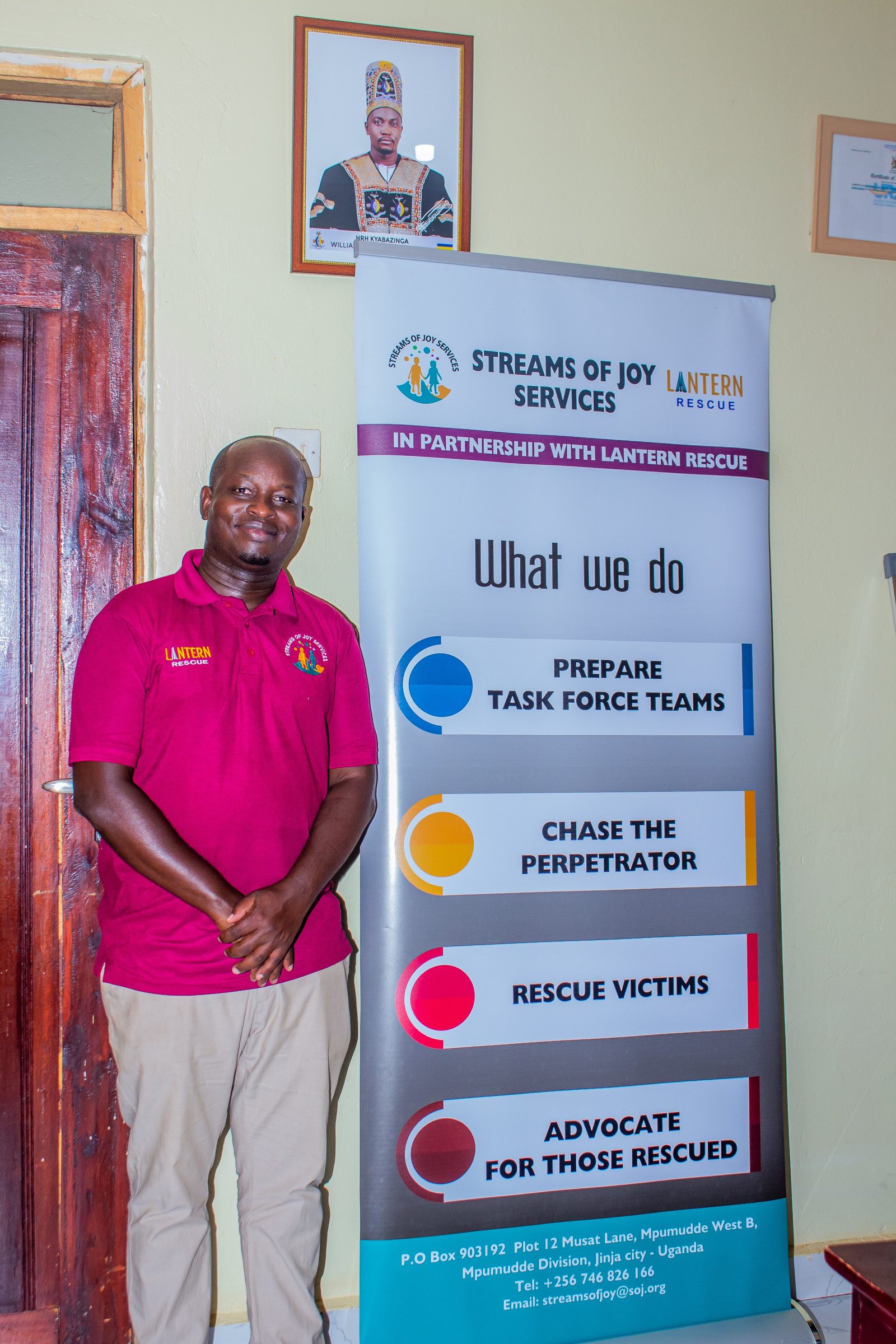Obed Byamugisha, founder of Streams of Joy Services, standing beside a banner setting out the organisation's work