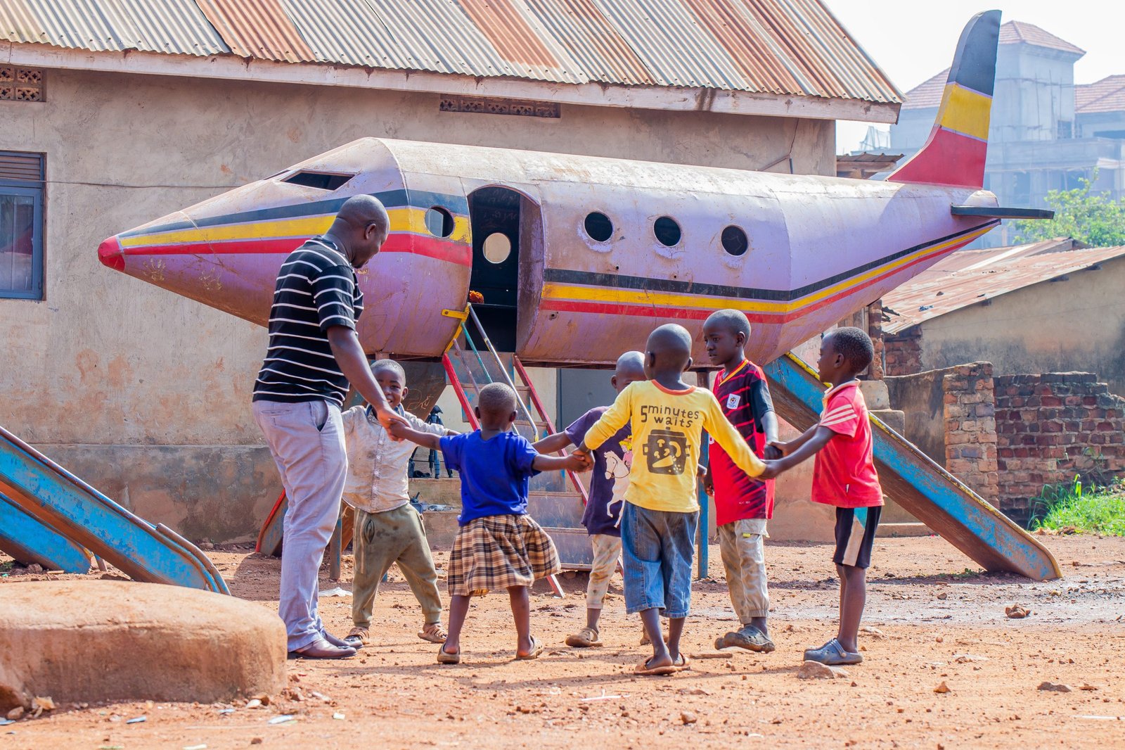 A Streams of Joy Services worker holding hands in a circle with children in front of a painted aeroplane playground in Uganda