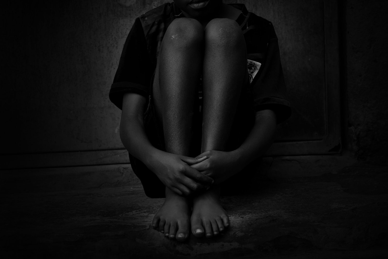 A child sitting alone against a wall in near darkness, barefoot, with their arms wrapped around their knees